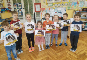 Uśmiechnie dzieci prezentują prace plastyczne przedstawiające jeża z dużymi kolcami wśród jesiennych liści.