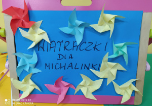 Zdjęcie przedstawia kolorowe papierowe wiatraczki przyklejone na niebieskim kartonie z napisem "wiatraczki dla Michalinki"
