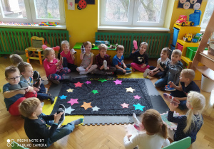 Zdjęcie przedstawia dzieci z grupy pszczółek siedzące na podłodze wokół czarnego zaczarowanego materiału na którym znajdują się kolorowe gwiazdy