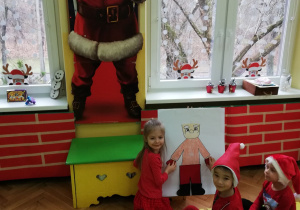 Zdjęcie przedstawia troje dzieci z grupy "Pszczółek" w czerwonych strojach oraz "Mikołajkowych" czapkach siedzących na podłodze przed tablicą z rysunkiem Mikołaja