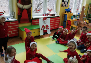 Zdjęcie przedstawia dzieci z grupy "Pszczółek" w czerwonych strojach oraz "Mikołajkowych" czapkach siedzących na podłodze w kole.