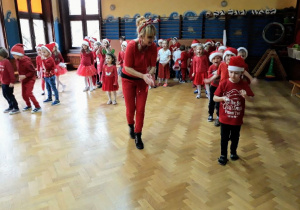 Radosne dzieci ubrane w czerwone czapki Mikołaj, uczestniczą w zabawie z okazji imienin Mikołaja. Prezentują swoje umiejętności taneczne wspólnie ze swoim nauczycielką.