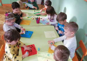 Na zdjęciu dzieci siedzą przy stolikach i ozdabiają kwadratowe, kolorowe kartki.