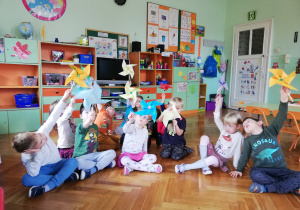 Na zdjęciu dzieci siedzą na podłodze, pokazują wykonane przez siebie kolorowe wiatraczki z papieru