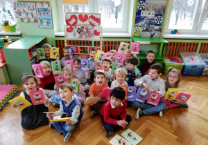 Na zdjęciu widać siedzące dzieci na podłodze, trzymające w rączkach wykonane przez siebie laurki dla babci i dziadka. W tle tablica z dekoracją z okazji Dnia babci i Dziadka