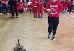 na zdjęciu chłopiec, który niesie bombkę choinkową na łyżce, w tle dzieci, które czekają na swoją kolej w konkursie, dzieci w strojach z dominującym kolorem czerwonym