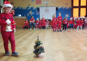 na zdjęciu chłopiec, który niesie bombkę choinkową na łyżce, w tle dzieci, które siedzą na ławeczce i dopingują chłopcu, dzieci w strojach z dominującym kolorem czerwonym