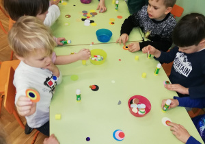 na zdjęciu dzieci siedzą przy stoliku, kleją pawie oczka z papieru. Na stole materiały potrzebne do wykonania pawich oczek: kółka w trzech wielkościach kolorowego papieru i kleje