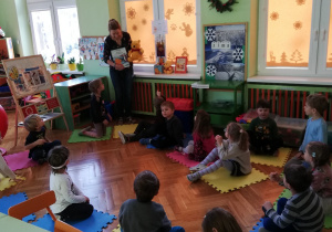 na zdjęciu - widok klasy przedszkolnej, dzieci siedzące na podłodze patrzą na nauczycielkę, która pokazuje książeczkę o Kubusiu Puchatku