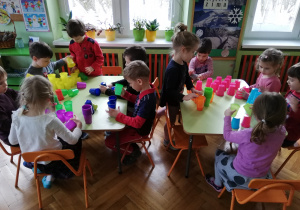 Na zdjęciu widać grupę dzieci w klasie przedszkolnej, przy stolikach, na których układają z kolorowych, plastikowych kubeczków wieże i inne budowle.