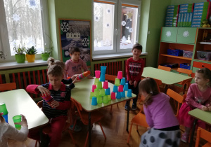 Na zdjęciu widać grupę dzieci w klasie przedszkolnej, przy stolikach, na których układają z kolorowych, plastikowych kubeczków wieże i inne budowle.
