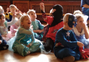 dzieci, w różnych strojach przebierańcowych siedzą na podłodze, patrzą z zaciekawieniem na coś poza planem zdjęcia