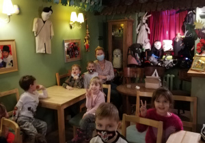 Na zdjęciu widok sali w " Baśniowej Kawiarence", w której siedzą dzieci na krzesełkach przy stolikach. W tle na ścianach widać maski i rekwizyty wykorzystywane w spektaklach granych przez aktorów Baśniowej Kawiarenki.