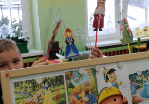 Na zdjęciu dzieci bawią się w teatr - na zdjęciu widać stojąca niską tablicę, na której przyczepione są różne obrazki z bajek, nad nią widać rączki dzieci trzymające papierowe kukiełki do bajki "O dzielnym Szewczyku".