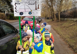 Na zdjęciu grupa dzieci stojących na chodniku, przy ulicy, na której stoi wiele samochodów. Dwoje dzieci na czele grupy trzyma transparent z napisem "Stop smogowi".