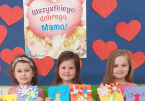 Na zdjęciu trzy dziewczynki pokazują wykonane przez siebie laurki, w tle serca i napis "Wszystkiego dobrego Mamo"