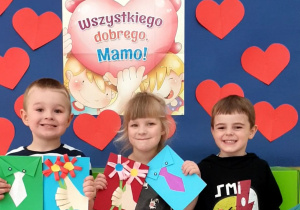Na zdjęciu troje dzieci pokazuje wykonane przez siebie laurki, w tle serca i napis "Wszystkiego dobrego Mamo"