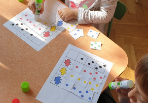 Na zdjęciu dzieci siedzące przy stoliku wykonujące kartę pracy - kodowanie z kropkami.