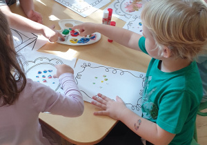 Na zdjęciu dzieci siedzące przy stoliku, wykonujące pracę plastyczną farbami.