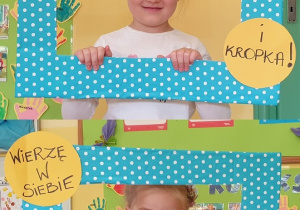 Na zdjęciu dwoje dzieci pozujących w niebieskiej ramce w kropki z napisem :Wierzę w siebie.. i kropka!"