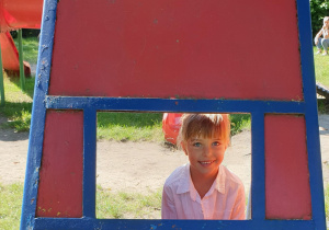 na zdjęciu dziewczynka wyglądająca przez okienko w domku na placu zabaw.