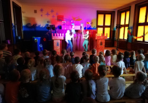 przedszkolaki oglądają występ aktorów, w tle scenografia do przedstawienia i aktorzy pokazujący duże kukiełki