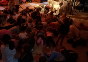 Dzieci kucają na podłodze, w tle prowadząca kierująca zabawą