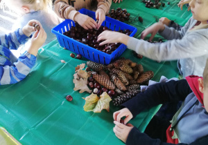 Na zdjęciu: w ogrodowej altance grupa dzieci segreguje kasztany, żołędzie , szyszki ułożone na stole figurki i ludziki z wykorzystaniem kasztanów, żołędzi, szyszek, patyczków
