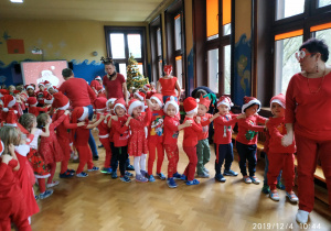 Dzieci ubrane w czerwone stroje oraz mikołajowe czapki tańczą ustawione jedno za drugim