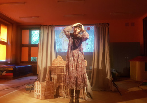 aktorka w brązowej wzorzystej sukience na tle sceny z makietą łódzkich kamienic