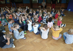 Dzieci siedzą na sali gimnastycznej, trzymają ręce na głowach