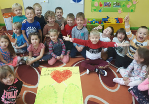 Na zdjęciu dzieci siedzące na dywanie radośnie i z uśmiechem przedstawiające efekty swojej grupowej pracy plastycznej.