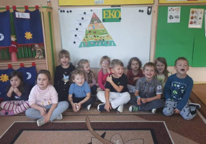 Dzieci z grupy trzeciej siedzą na dywanie, w tle na tablicy umieszczona jest Piramida żywienia i żółty napis Eko na zielonym tle.