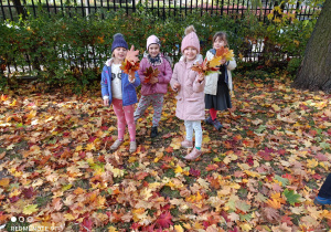 Na zdjęciu stoją dziewczynki w ogrodzie przedszkolnym, trzymając w rękach bukiety z kolorowych liści.