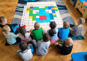 Grupa dzieci siedzących tyłem na dywanie w siadzie skrzyżnym. Przygląda sie obrazkowi na macie. Wykonane zadanie przedstawia zakodowaną głowę misia, wykonanego z kolorowych kwadratów.