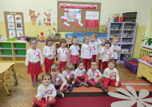 Dzieci z grupy pierwszej pozują w biało czerwonych strojach w klasie na tle kącika patriotycznego