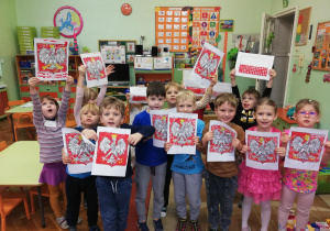 Grupa dzieci pokazuje własne prace plastyczne " Godło polski" i Flaga Polski" -wykonane z użyciem farb