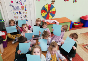 Dzieci z niebieskimi kartkami w rękach siedzą pod tablicą z Prawami Dziecka