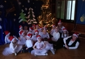 Dzieci siedzą pod przedszkolną choinka w pięknych strojach świątecznych.
