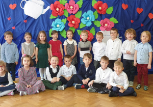 Zdjęcie przedstawia grupę dzieci w odświętnych ubraniach na tle dekoracji z kolorowych kwiatów.
