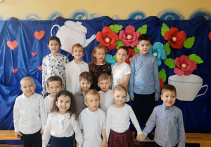 Grupa dzieci przedszkolnych ubranych w stroje galowe, pozuje do zdjęcia na tle dekoracji z kolorowych kwiatów przygotowanej z okazji Dnia Babci i Dziadka.