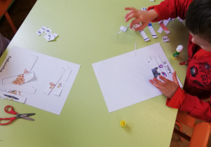 Dzieci przy stolikach wycinają puzzle i naklejają w odpowiednim ułożeniu na kartce