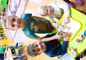 Na zdjęciu trójka dzieci bawi się mąką, dotykając jej.