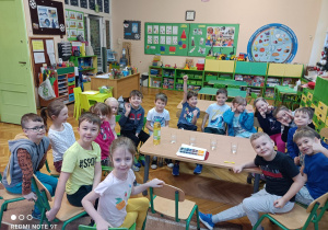 Na zdjęciu grupa dzieci siedzi na krzesełkach obserwują materiały potrzebne do przeprowadzenia eksperymentu wody z olejem i kolorowymi kostkami lodu.