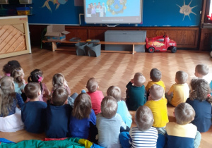 Na zdjęciu dzieci siedzące na podłodze na sali gimnastycznej oglądające film edukacyjny.