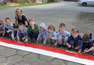 Na zdjęciu dzieci kucające przy malowanej fladze Polski wraz z nauczycielką.