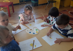 na zdjęciu dzieci siedzące przy stolikach w klasie wykonujące pracę plastyczną z wykorzystaniem farb plakatowych.