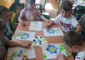 na zdjęciu dzieci siedzące przy stolikach w klasie wykonujące pracę plastyczną z wykorzystaniem farb plakatowych.