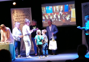 Nauczycielka wraz z dziećmi odbiera nagrodę za udział w konkursie " Przedszkolne orkiestry" - widok sceny, na której stoją dzieci z nauczycielka i jury
