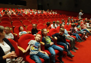 Na zdjęciu dzieci siedzące na czerwonych fotelach w sali teatralnej.